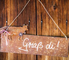 Metal sign with a cow figure, arrow and the words “Griaß di!” hanging on a wooden wall. | © SalzburgerLand Tourismus, Achim Meurer