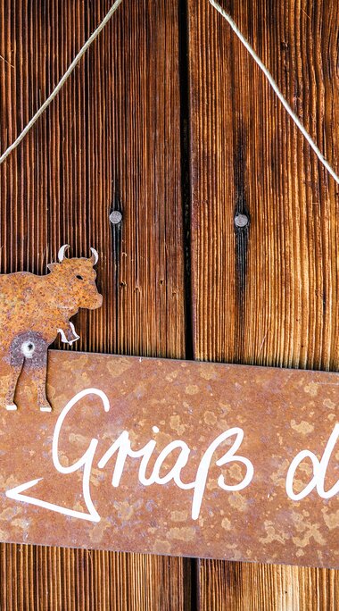 Metallschild mit Kuhfigur, Pfeil und der Aufschrift „Griaß di!“ an einer Holzwand. | © SalzburgerLand Tourismus, Achim Meurer