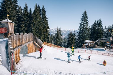 Erwachsene und Kinder fahren auf Ski durch die Funslope Schmitten mit Holzelementen und Drachenmaskottchen. | © Zell am See-Kaprun Tourismus