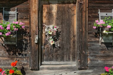 Alte Holztür in Niedernsill mit Blumenschmuck und bepflanzten Holzkästen an einer dunklen Holzfassade. | © TVB Piesendorf Niedernsill, Harry Liebmann