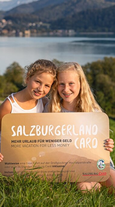 Two children hold the SalzburgerLand Card on a meadow, with a lake and mountains in the background. | © Michael Groessinger