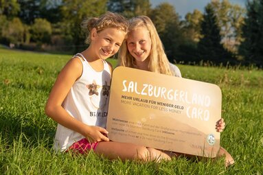 Two children sit on a meadow and hold a large SalzburgerLand Card towards the camera. | © Michael Groessinger