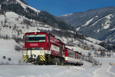 Pinzgauer Lokalbahn travels through a snowy valley with houses and forested mountain slopes. | © Salzburg AG