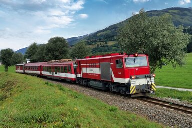 Red and white Pinzgauer Lokalbahn travelling between green meadows and forested mountain slopes. | © Florian Knapp