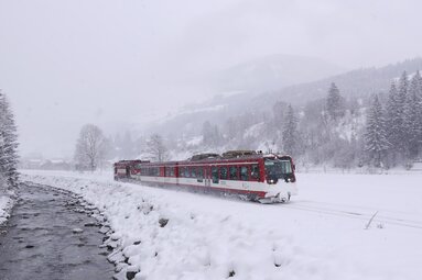 Pinzgauer Lokalbahn fährt bei starkem Schneefall entlang eines Bachs durch eine verschneite Winterlandschaft. | © Florian Knapp