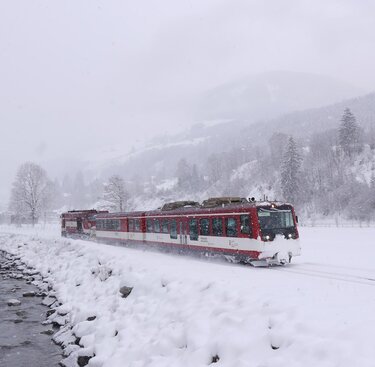 Pinzgauer Lokalbahn travels beside a stream through a snow-covered landscape during heavy snowfall. | © Florian Knapp
