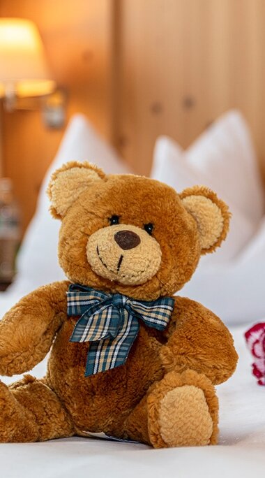 Teddy bear with a checked bow on a white-covered bed at Gästehaus Klawunn, with a red and white flower beside it. | © nikolaus faistauer photography