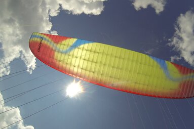 Farbiger Gleitschirm von unten vor blauem Himmel mit Wolken und sichtbarer Sonne. | © sky-42.com