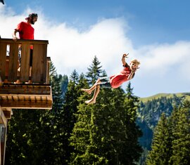 Ein Junge springt von einer Holzplattform, während ein Mann zusieht. Im Hintergrund sind Wald und Berge zu sehen. | © saalbach.com, Foto Mirja Geh