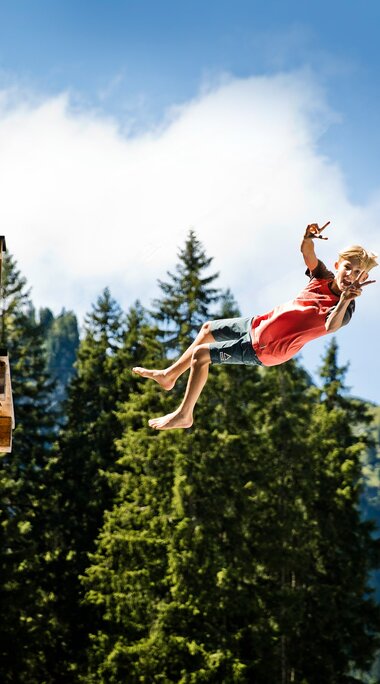 A boy jumps from a wooden platform while a man watches. Forest and mountains are visible in the background. | © saalbach.com, Foto Mirja Geh