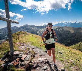 rail runner passes a wooden cross on a mountain trail, with an alpine mountain landscape in the background. | © SalzburgerLand Tourismus © Foto Philipp Freund