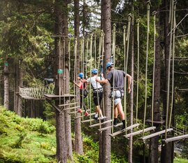 Three people wearing helmets and climbing harnesses cross suspended wooden planks at a high ropes course in a forest. | © saalbach.com, Foto Mia Knoll