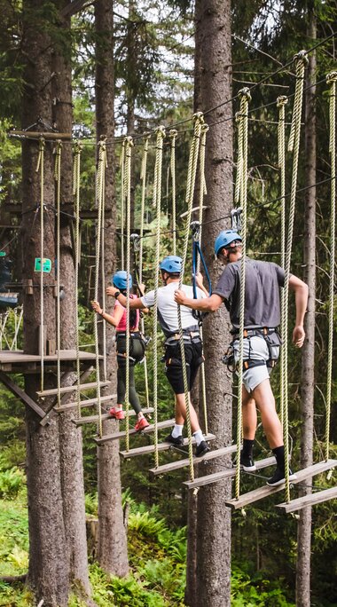 Drei Personen mit Helm und Klettergurt überqueren frei hängende Holzbretter in einem Hochseilpark im Wald. | © saalbach.com, Foto Mia Knoll