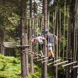 Three people wearing helmets and climbing harnesses cross suspended wooden planks at a high ropes course in a forest. | © saalbach.com, Foto Mia Knoll