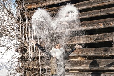 Person throws snow into the air in front of an old wooden hut, with icicles and a snowy landscape in the background. | © TVB Piesendorf Niedernsill, Foto Harry Liebmann