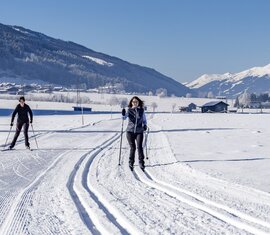 Two people cross-country skiing on groomed trails with snow-covered mountains in the background. | © TVB Piesendorf Niedernsill, Foto Lichtfarben