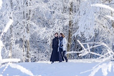 Two people walking together through a snowy forest with snow-covered trees. | © Lichtfarben