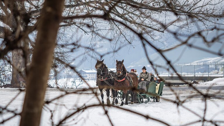 Zwei Pferde vor einem Schlitten mit Fahrgästen auf einer verschneiten Fläche vor winterlichen Berghängen. | © TVB Piesendorf Niedernsill, Foto Lichtfarben 