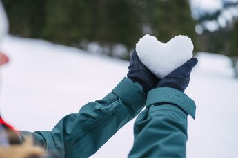 Person in Winterkleidung hält ein Herz aus Schnee in den Händen, im Hintergrund eine verschneite Landschaft. | © NATURESA, Fotograf Christoph Oberschneider 
