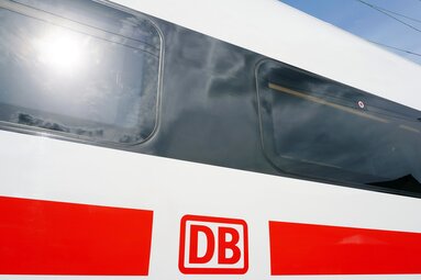 Close-up of a red and white Deutsche Bahn train with DB logo and reflective windows beneath overhead wires. | © Deutsche Bahn AG / Volker Emersleben