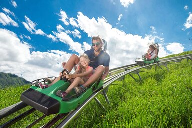 Erwachsener und Kind fahren gemeinsam mit einer Alpen-Achterbahn über einen grünen Berghang. | © Kitzsteinhorn