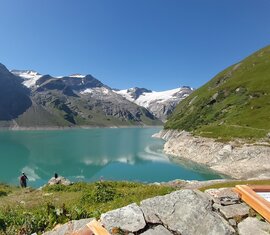 Hochgebirgsstausee bei Kaprun mit zwei Personen am Ufer, umgeben von grünen Berghängen und schneebedeckten Gipfeln. | © TVB Piesendorf Niedernsill