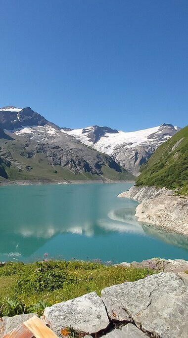 Hochgebirgsstausee bei Kaprun mit zwei Personen am Ufer, umgeben von grünen Berghängen und schneebedeckten Gipfeln. | © TVB Piesendorf Niedernsill