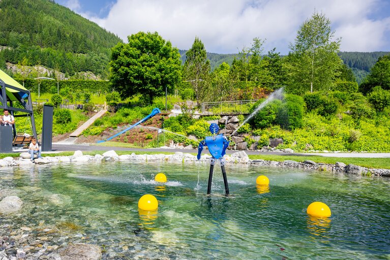 Krimmler WasserWelten mit Wasserbecken, Fontänen, Spielelementen und Rutsche vor bewaldeten Berghängen. | © krimmler-wasserwelten.at / Stabentheiner