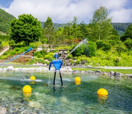 Krimml Water Worlds with a water basin, fountains, play features and a slide in front of forested mountain slopes. | © krimmler-wasserwelten.at / Stabentheiner
