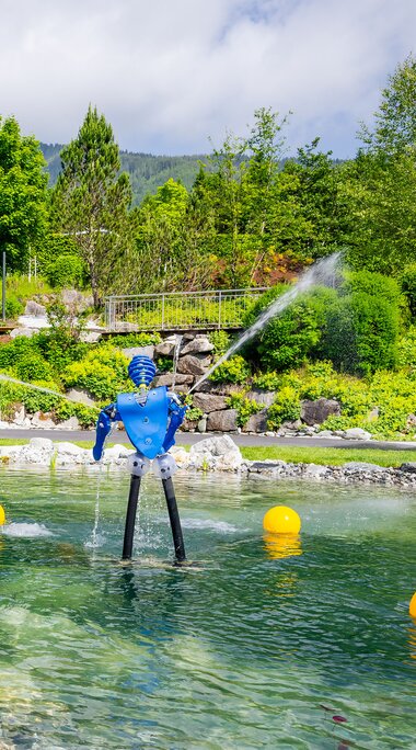 Krimml Water Worlds with a water basin, fountains, play features and a slide in front of forested mountain slopes. | © krimmler-wasserwelten.at / Stabentheiner