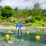 Krimml Water Worlds with a water basin, fountains, play features and a slide in front of forested mountain slopes. | © krimmler-wasserwelten.at / Stabentheiner