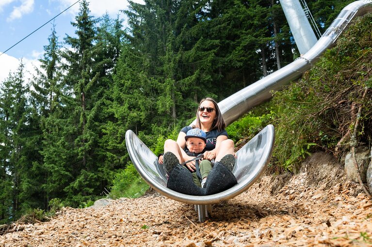 Erwachsene Person und Kind auf dem Rutschenweg der Wildkogel-Arena, daneben eine Gondelbahn und Wald. | © Wildkogel Arena Neukirchen & Bramberg