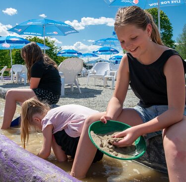 Kinder spielen im Edelsteinpark mit Sand und Wasser an einer flachen Wasserrinne vor alpiner Bergkulisse. | © Edith Danzer