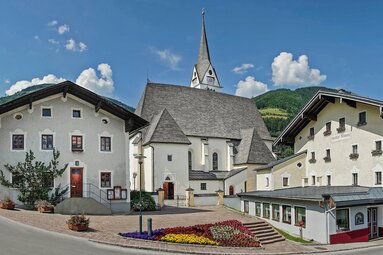 Piesendorf parish church with its steeple and surrounding houses, flower beds in front and wooded mountain slopes behind. | © TVB Piesendorf Niedernsill, Foto Harry Liebmann