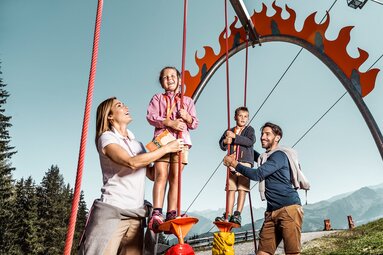 Zwei Kinder balancieren auf Spielelementen an Seilen, begleitet von zwei Erwachsenen vor einer Bergkulisse. | © Schmittenhöhebahn AG