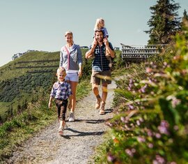 Familie mit zwei Kindern beim Wandern auf einem Bergweg der Schmittenhöhe, im Hintergrund schneebedeckte Gipfel. | © Schmittenhöhebahn AG