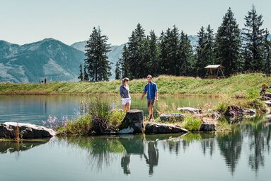 Two hikers at the Plettsaukopf reservoir, surrounded by meadows, conifers and mountains. | © Schmittenhöhebahn AG