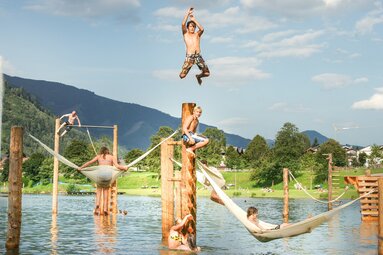 Kinder und Jugendliche klettern und spielen an Holzelementen und Hängematten im Wasserpark eines Badesees. | © TVB Piesendorf Niedernsill, Foto Harry Liebmann