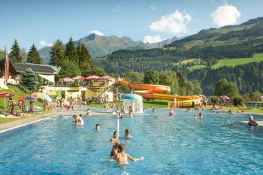Kinder und Erwachsene baden im Freibad mit Wasserrutsche vor bewaldeten Bergen. | © TVB Piesendorf Niedernsill, Foto Harry Liebmann