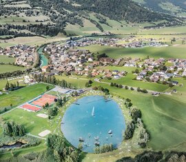 Aerial view of Niedernsill swimming lake with lawns and sports courts, with the village and green mountain slopes behind. | © TVB Piesendorf Niedernsill, Foto Harry Liebmann