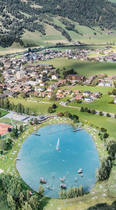 Luftaufnahme des Badesees Niedernsill mit Liegewiesen und Sportplätzen, dahinter der Ort und grüne Berghänge. | © TVB Piesendorf Niedernsill, Foto Harry Liebmann