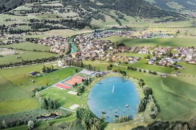 Luftaufnahme des Badesees Niedernsill mit Liegewiesen und Sportplätzen, dahinter der Ort und grüne Berghänge. | © TVB Piesendorf Niedernsill, Foto Harry Liebmann