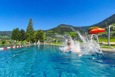 Person springt ins Becken im Hinkelsteinbad Piesendorf, im Hintergrund liegen Liegewiese und Berge. | © Gemeinde Piesendorf, Foto Lichtfarben