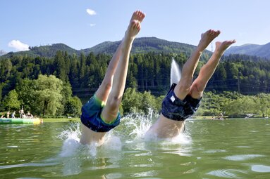 Zwei Personen springen kopfüber in den Badesee Niedernsill, im Hintergrund sind Badegäste und bewaldete Berge zu sehen. | © Ferienregion Nationalpark Hohe Tauern, Foto Michael Huber