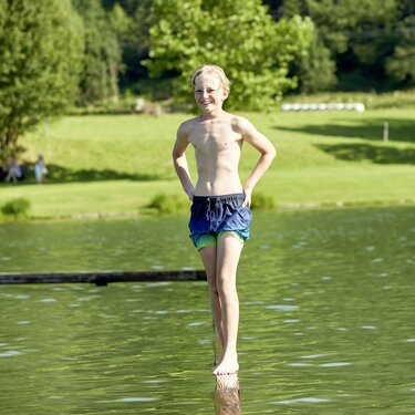 Child standing on a wooden post in Badesee Niedernsill, with a lawn and trees in the background. | © Ferienregion Nationalpark Hohe Tauern, Foto Michael Huber