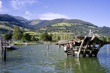 Badende am Badesee Niedernsill mit hölzerner Spielanlage im Wasser und grünen Berghängen im Hintergrund. | © Ferienregion Nationalpark Hohe Tauern, Foto Michael Huber
