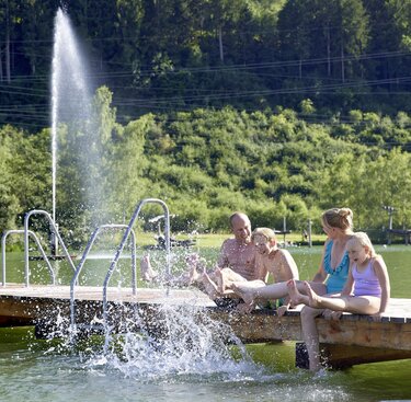 Familie sitzt auf einem Holzsteg am Badesee Niedernsill und spielt gemeinsam mit den Füßen im Wasser. | © Ferienregion Nationalpark Hohe Tauern, Foto Michael Huber
