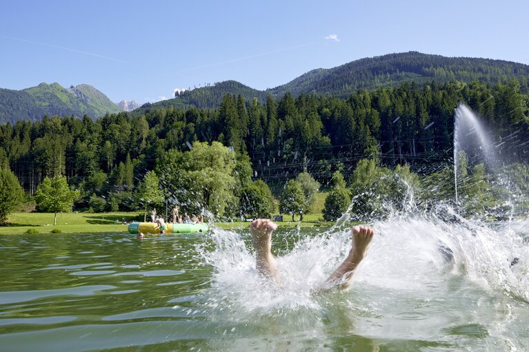 Person jumps into Badesee Niedernsill, with bathers, meadows, forest and mountains in the background. | © Ferienregion Nationalpark Hohe Tauern, Foto Michael Huber