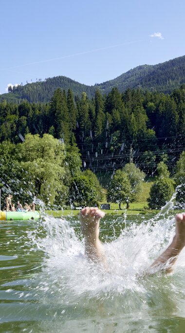 Person jumps into Badesee Niedernsill, with bathers, meadows, forest and mountains in the background. | © Ferienregion Nationalpark Hohe Tauern, Foto Michael Huber