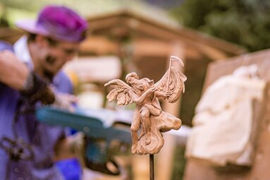 Geschnitzte Engelsfigur aus Holz mit Sense, im Hintergrund arbeitet ein Schnitzer beim Bartlmä Fest. | © Lichtfarben